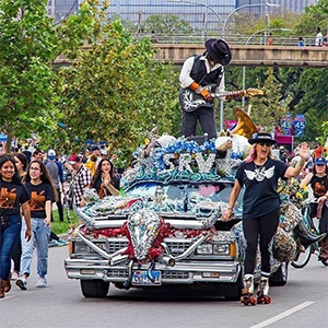 Man playing guitar while standing in a convertable art car in a parade, a woman roller skates along side the car