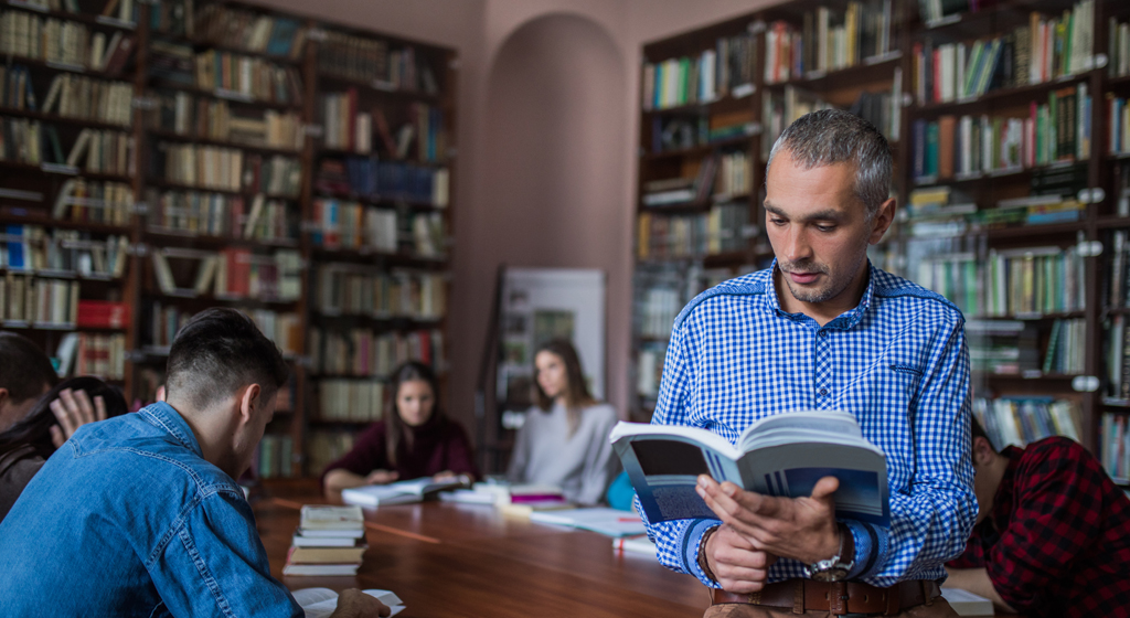 students reading at a large table in a room of books, man reading an open book facing forward.