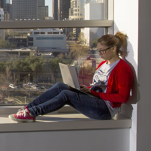 UHD student on a laptop in sitting in a window overlooking downtown Houston