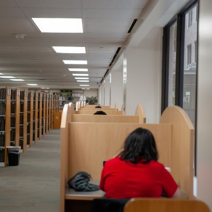 The shelves at the W.I. Dykes library on the left. On the right are study carrels. In the foreground, a student is studying in a carrel with her back to the camera. She is wearing a red shirt.