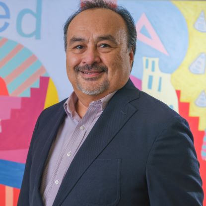 Henry Gonzales wears a blazer and is posing in front of a Floyd Newsum painting at the College of Public Service.