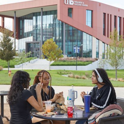 UHD Wellness & Success Center in the background. Three female students eat at a round table outdoors.