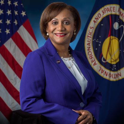 Vanessa E. Wyche posing for a professional headshot. She is in a bright blue suit. American and NASA flags are behind her.