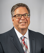 Dr. Daniel Maxwell poses for a professional headshot. He is wearing a charcoal suit with a checkered red tie.