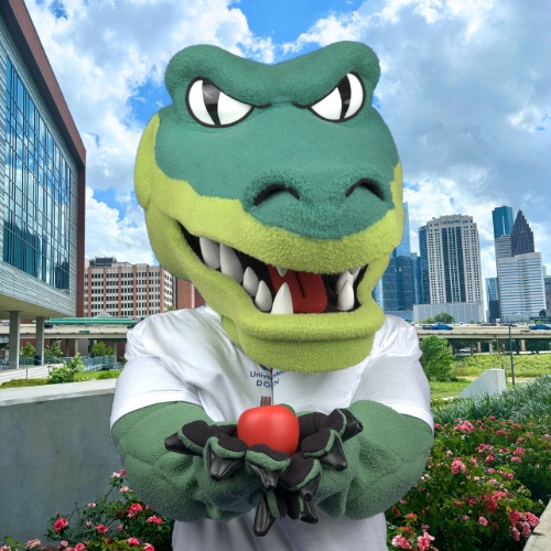 Ed-U-Gator holds an apple in front of the college of sciences and technology at the university of houston downtown